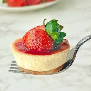 A fork holds a small cheesecake topped with glossy strawberry sauce and a fresh strawberry. The background is a soft focus, white marble surface.