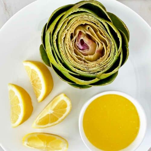 A plate with a halved artichoke, four lemon wedges, and a small bowl of yellow dipping sauce. A striped cloth is visible in the background.